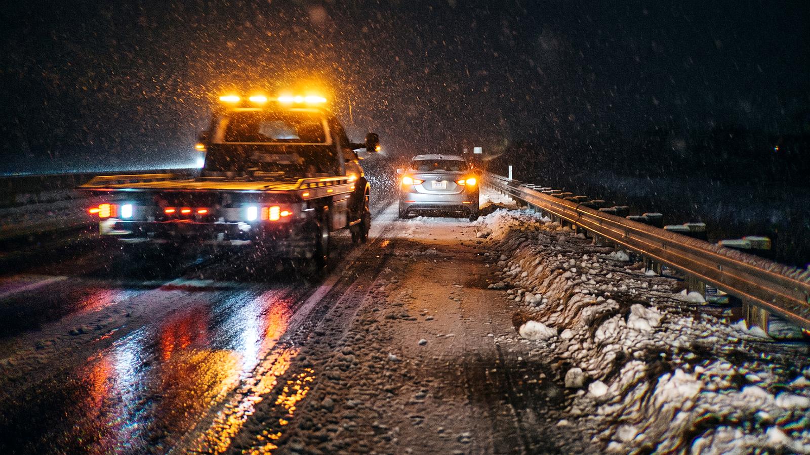 A tow truck's amber light bar on a snowy highway shoulder at night, a stopped car's lights ahead through falling snow