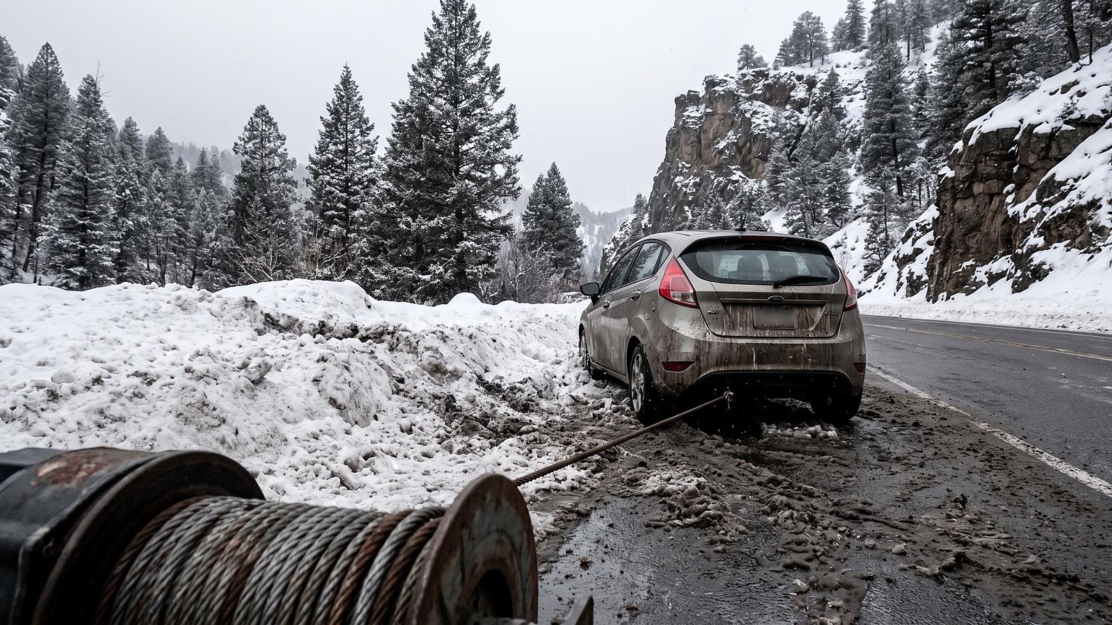 A car nosed into a plowed snowbank on a canyon road, a tow truck's winch spool and cable in the foreground